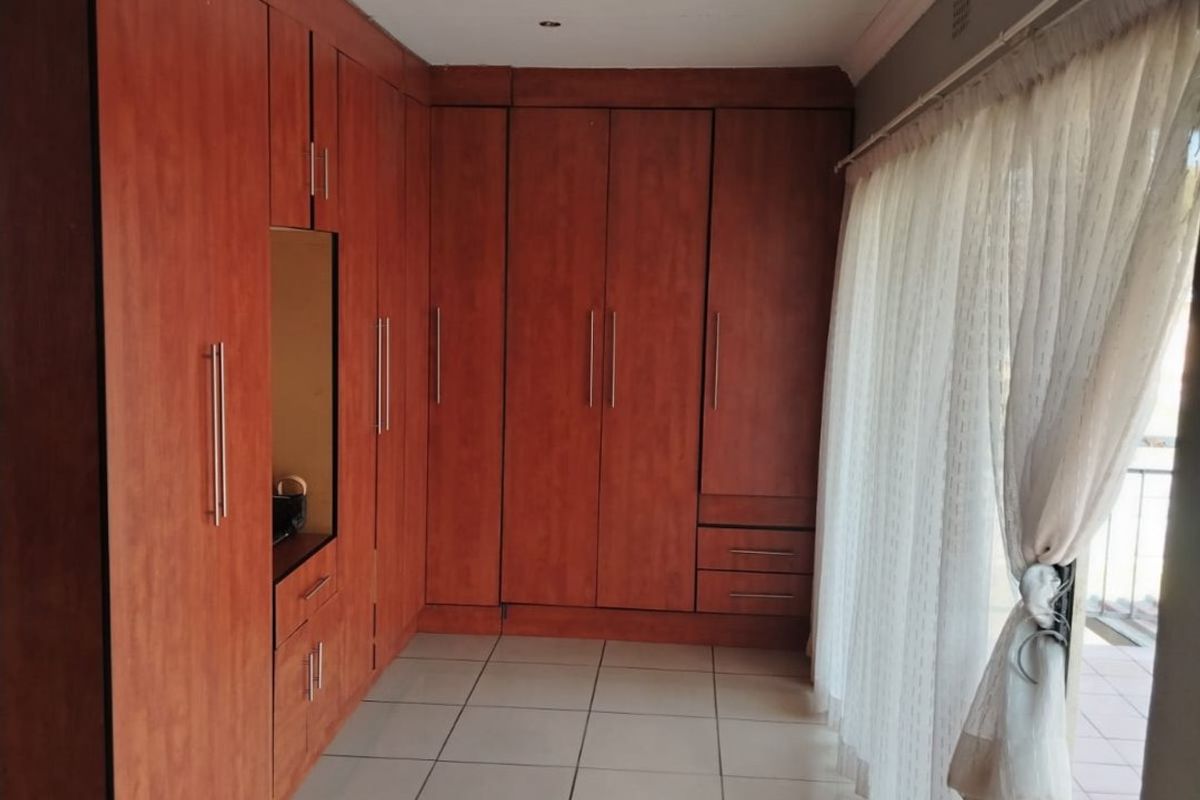 A bright bedroom featuring extensive built-in wooden cupboards, neutral tiled flooring, and a sliding glass door providing access to an outdoor area.