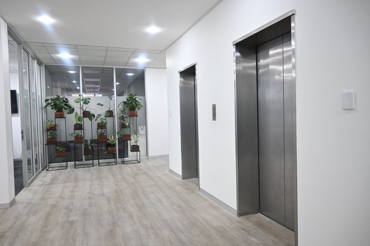 A clean and modern commercial hallway featuring stainless steel elevator access, glass partitions, and neutral laminate flooring with recessed lighting.