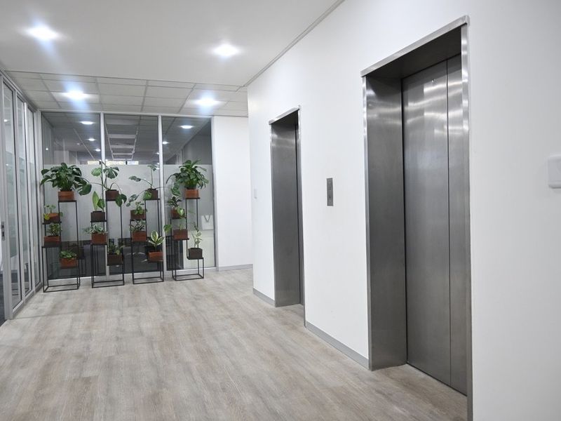A clean and modern commercial hallway featuring stainless steel elevator access, glass partitions, and neutral laminate flooring with recessed lighting.