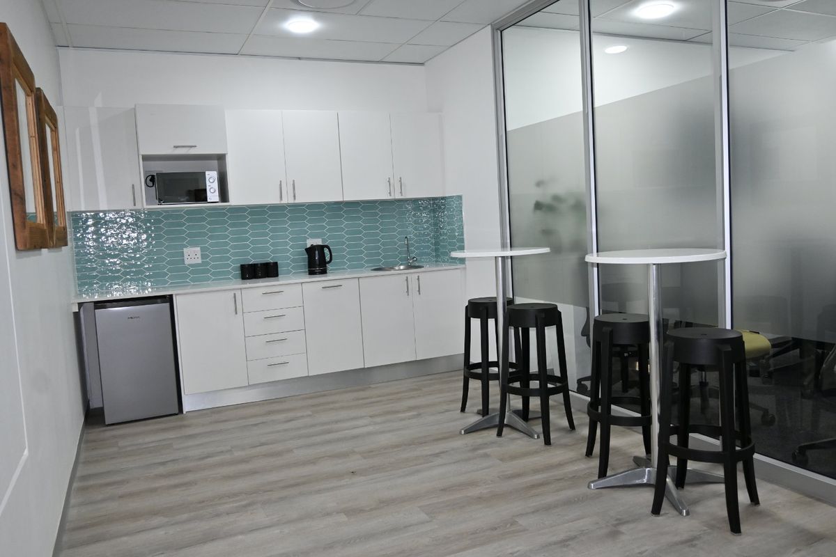 A modern office kitchenette featuring white cabinetry, a teal tiled backsplash, stainless steel sink, and a glass partition wall.