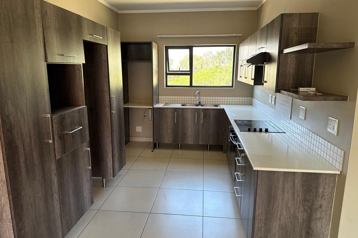 A modern, empty kitchen featuring dark wood-look cabinetry, white countertops, a built-in electric hob, and a stainless steel sink with a window view.