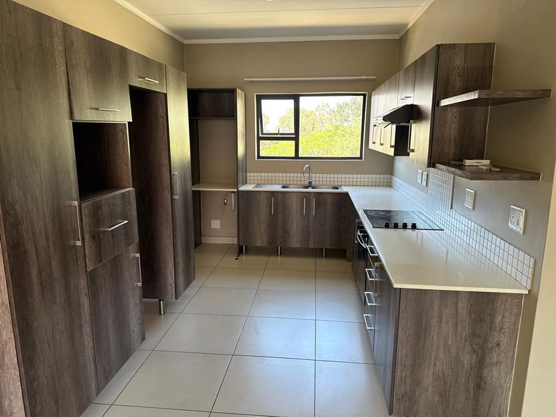 A modern, empty kitchen featuring dark wood-look cabinetry, white countertops, a built-in electric hob, and a stainless steel sink with a window view.