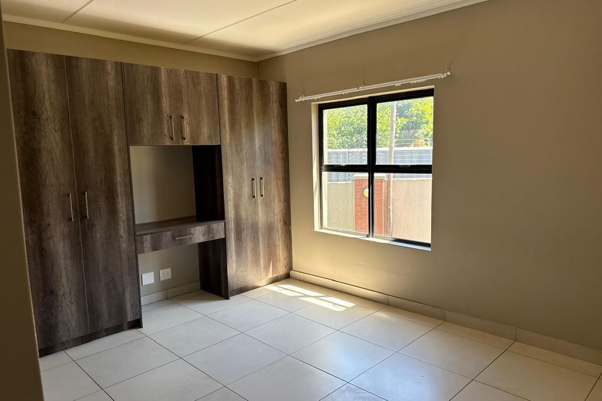 A bright, empty bedroom featuring neutral tiled flooring, a large window, and extensive built-in wooden cabinetry with an integrated desk area.
