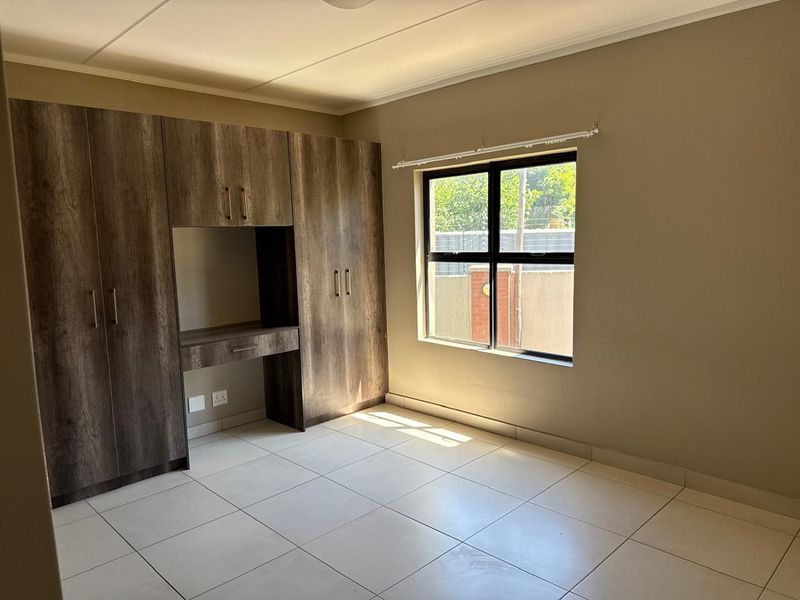 A bright, empty bedroom featuring neutral tiled flooring, a large window, and extensive built-in wooden cabinetry with an integrated desk area.