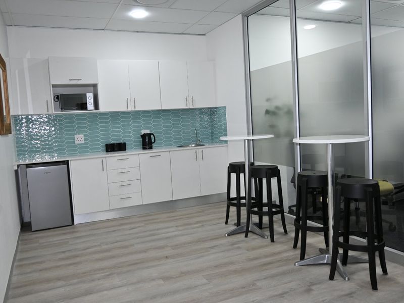 A modern office kitchenette featuring white cabinetry, a teal tiled backsplash, stainless steel sink, and a glass partition wall.