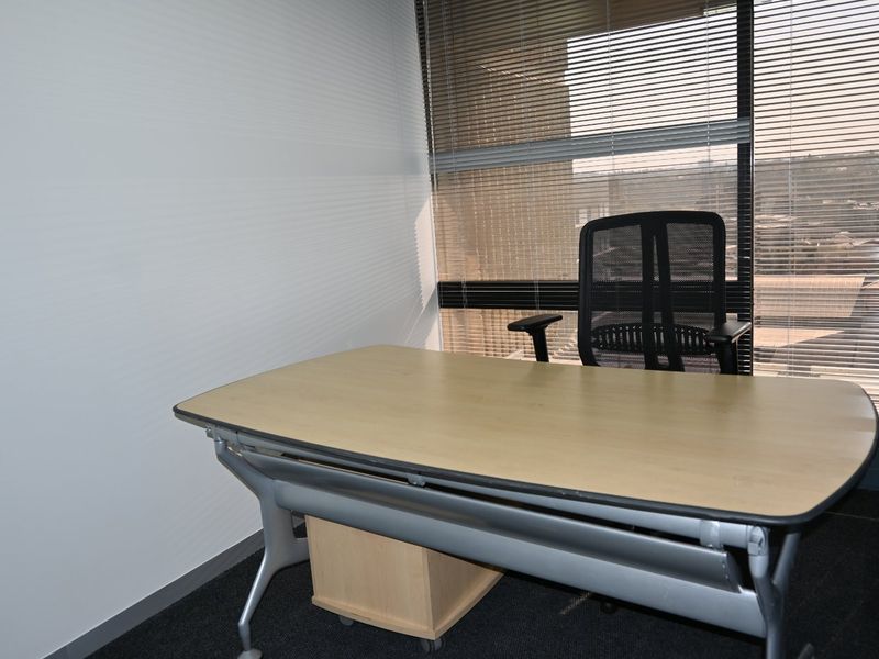 A functional commercial office space featuring carpet flooring and large windows with adjustable venetian blinds, providing natural light and an exterior view.