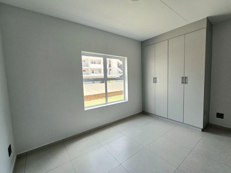 A bright, modern bedroom featuring neutral tiled flooring, built-in cupboards, and a large window providing natural light.