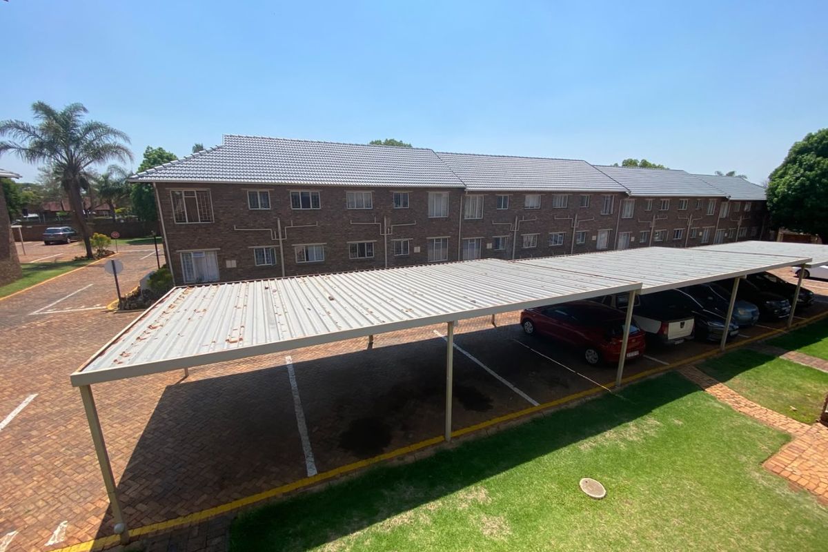 Exterior view of an apartment complex featuring a brick facade, tiled roof, and a covered carport parking area with paved driveway and lawn.