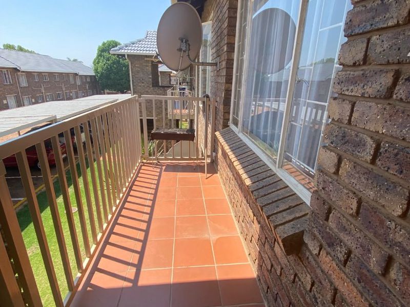 A functional balcony featuring tiled flooring, a metal balustrade, and exposed brick exterior walling, overlooking the apartment complex grounds.