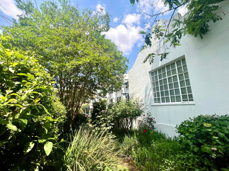 A view of the rear exterior facade featuring a white stucco finish and a classic grid-pattern window, surrounded by established garden greenery.
