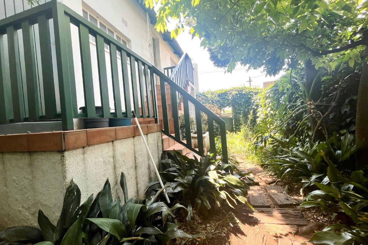 A rear garden view featuring a raised patio with a timber balustrade, staircase, and a garden path leading through established greenery.