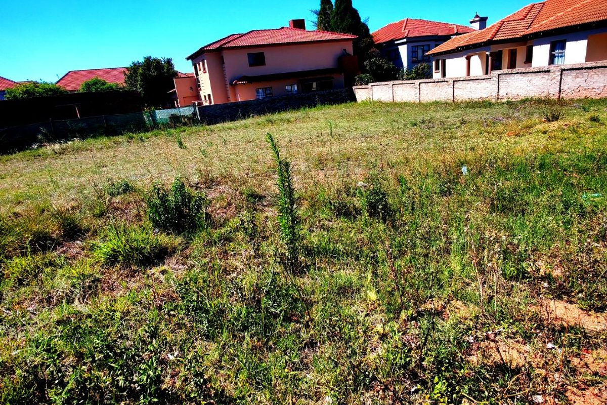 A vacant residential plot of land featuring natural grass coverage and a boundary wall, situated in a developed suburban neighborhood.