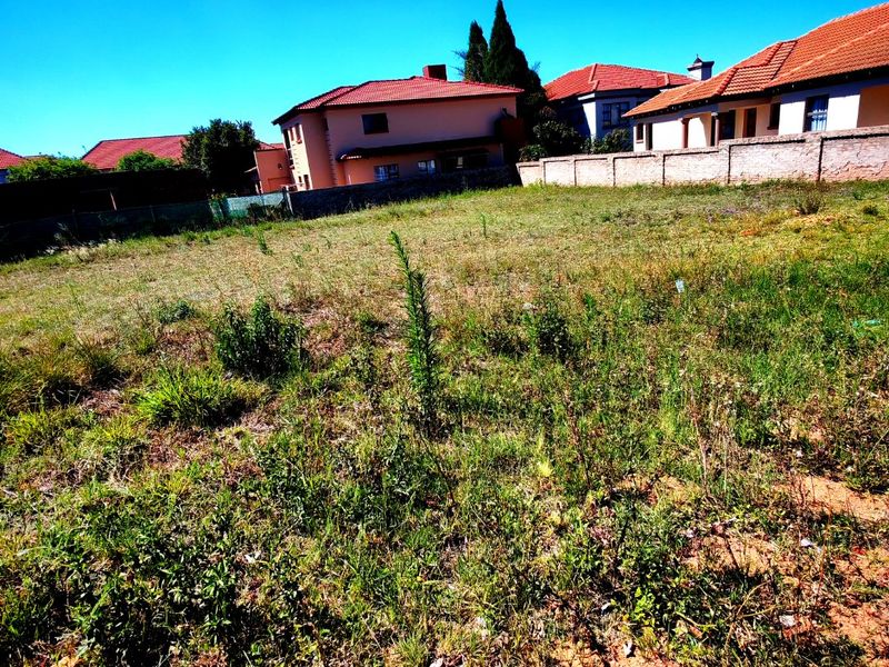 A vacant residential plot of land featuring natural grass coverage and a boundary wall, situated in a developed suburban neighborhood.