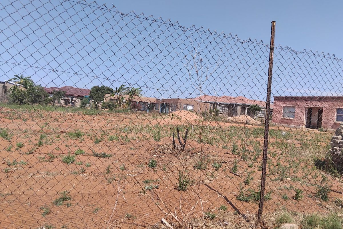 A view of a vacant residential plot enclosed by chain-link fencing, showing undeveloped land with surrounding neighborhood houses in the background.