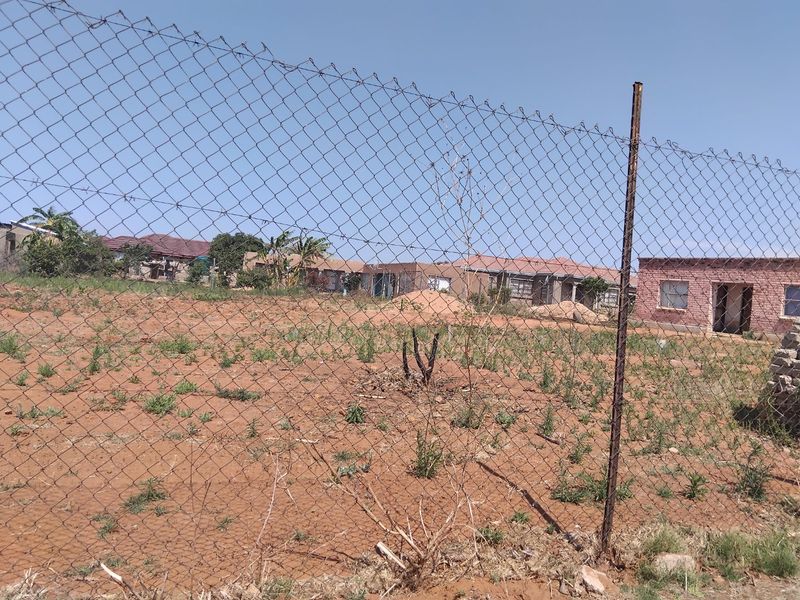 A view of a vacant residential plot enclosed by chain-link fencing, showing undeveloped land with surrounding neighborhood houses in the background.