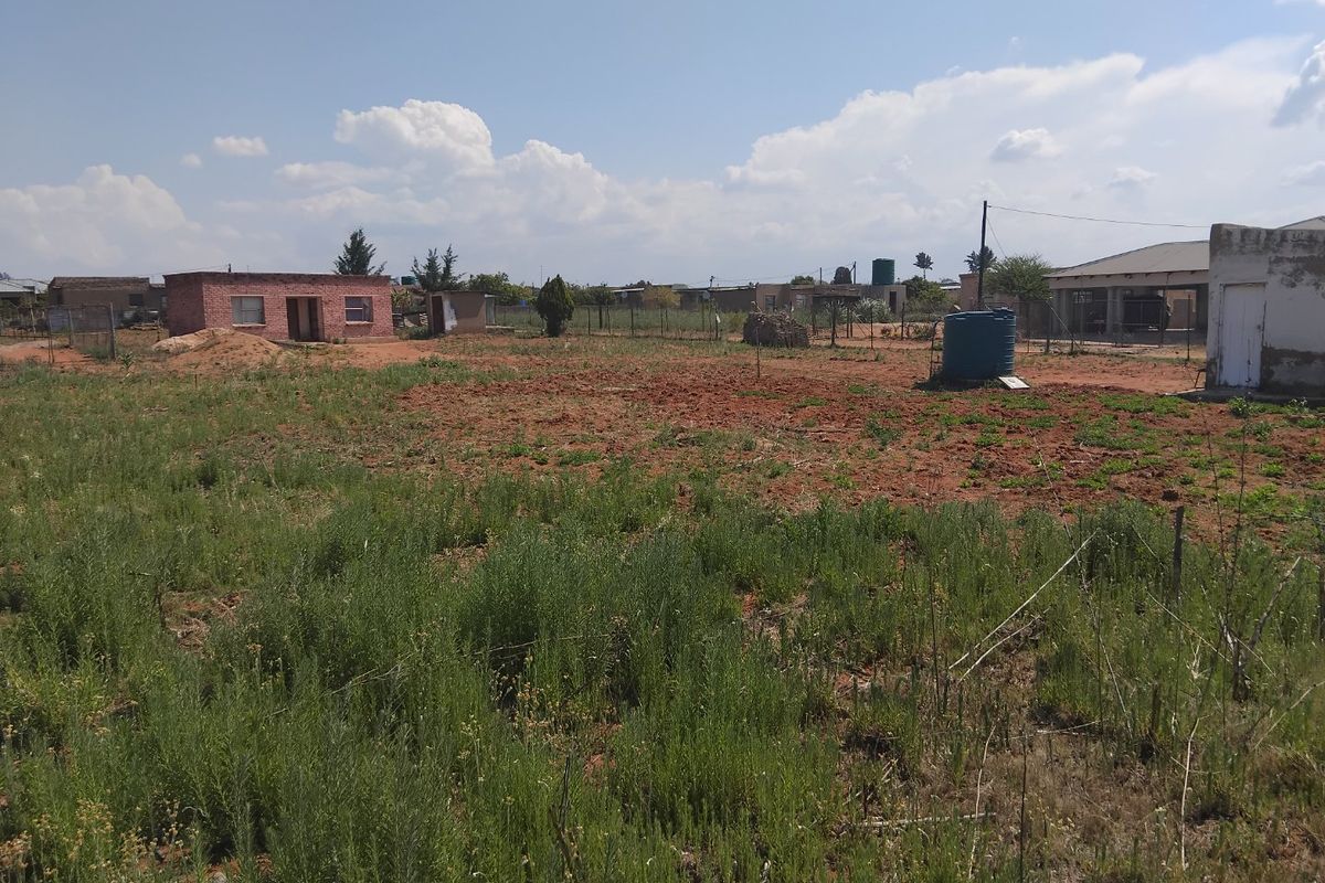 A view of a vacant residential plot featuring natural ground cover, perimeter fencing, and an existing outbuilding structure.