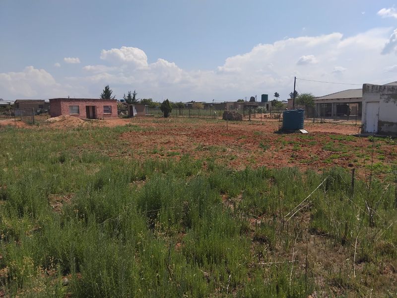 A view of a vacant residential plot featuring natural ground cover, perimeter fencing, and an existing outbuilding structure.