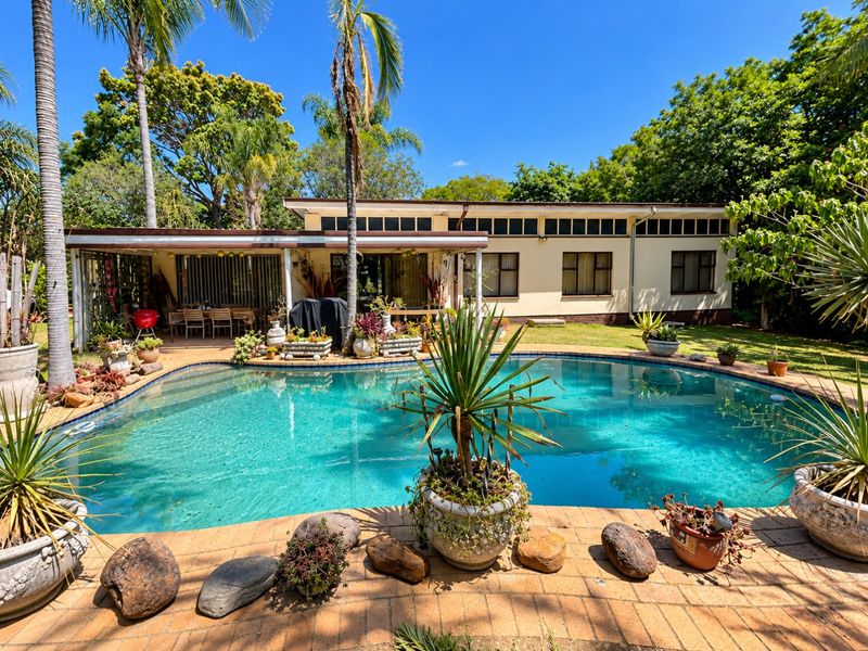 A well-maintained outdoor pool area featuring a paved deck, a covered patio space, and established garden surroundings, perfect for outdoor relaxation and entertainment.