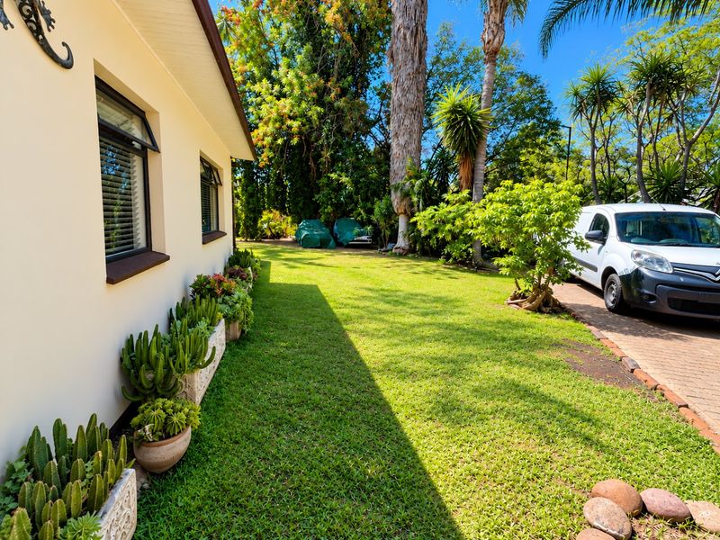 A well-maintained side garden featuring a lush green lawn, established trees, and a paved driveway area alongside the house exterior.