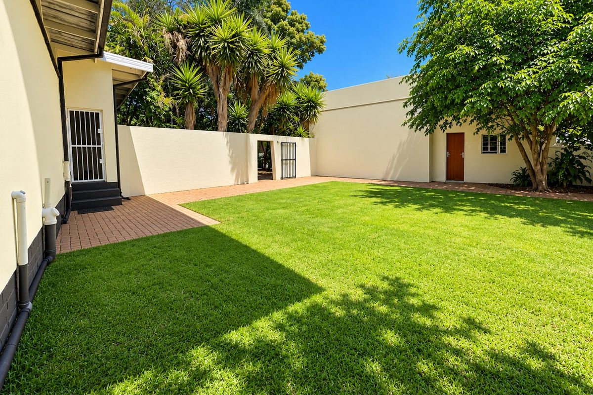 A well-maintained rear garden featuring a paved patio area, lush green lawn, and secure boundary walls with gated access.
