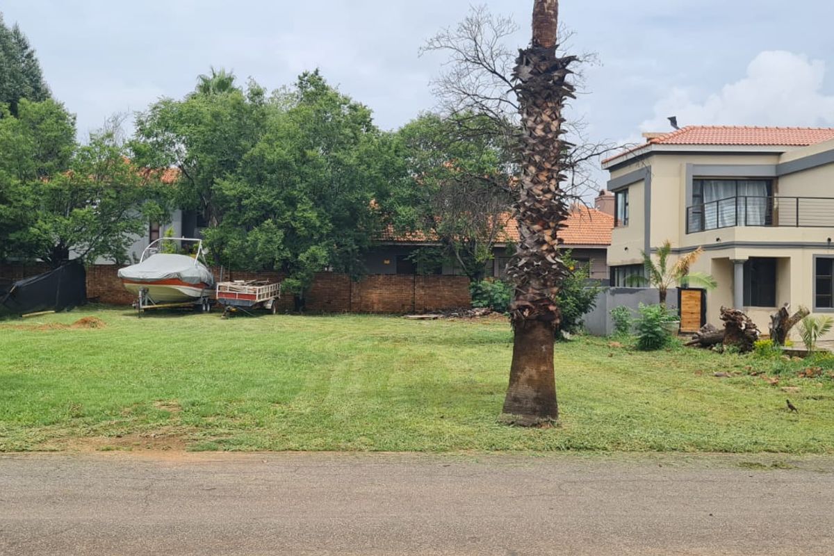 A vacant residential plot featuring a well-maintained lawn, mature palm trees, and a brick boundary wall, situated adjacent to an existing residential property.