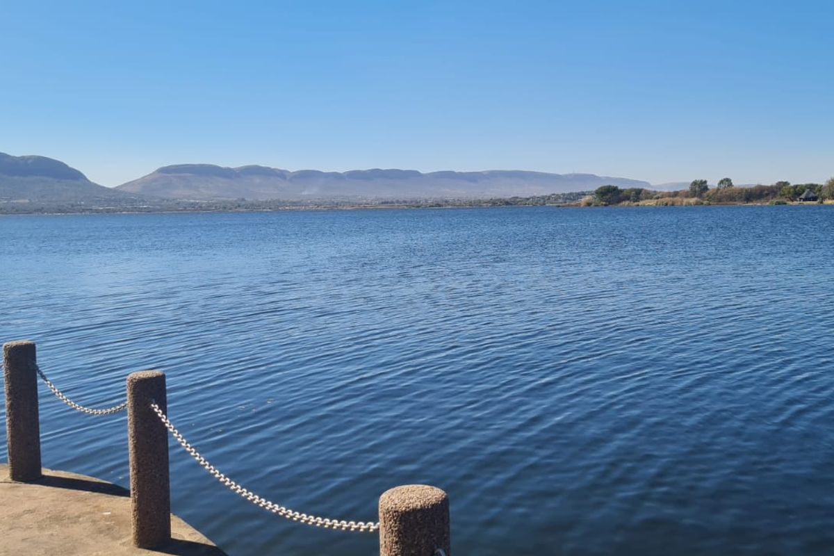 A scenic view overlooking a large lake with distant mountains, featuring a concrete pier edge with decorative bollards and chain railing.