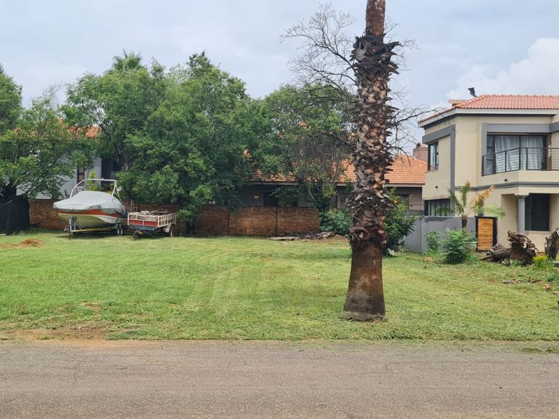 A vacant residential plot featuring a well-maintained lawn, mature palm trees, and a brick boundary wall, situated adjacent to an existing residential property.