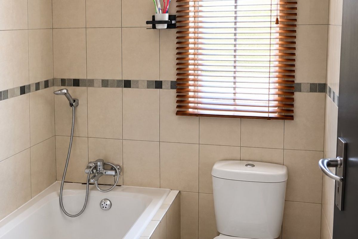 A clean, functional bathroom featuring neutral wall tiles, a built-in bathtub with a handheld shower mixer, and a standard toilet.