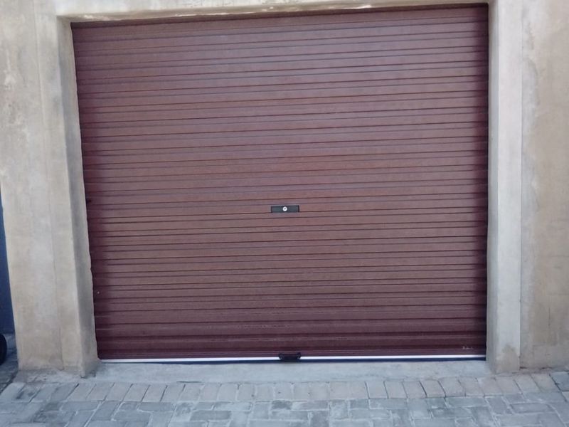 A single garage featuring a brown roller shutter door set within an unfinished concrete facade and a paved driveway.
