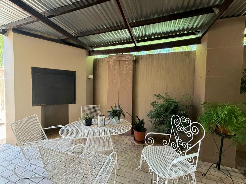 Covered patio area featuring a built-in braai, paved flooring, and a corrugated metal roof structure, providing a functional outdoor entertainment space.