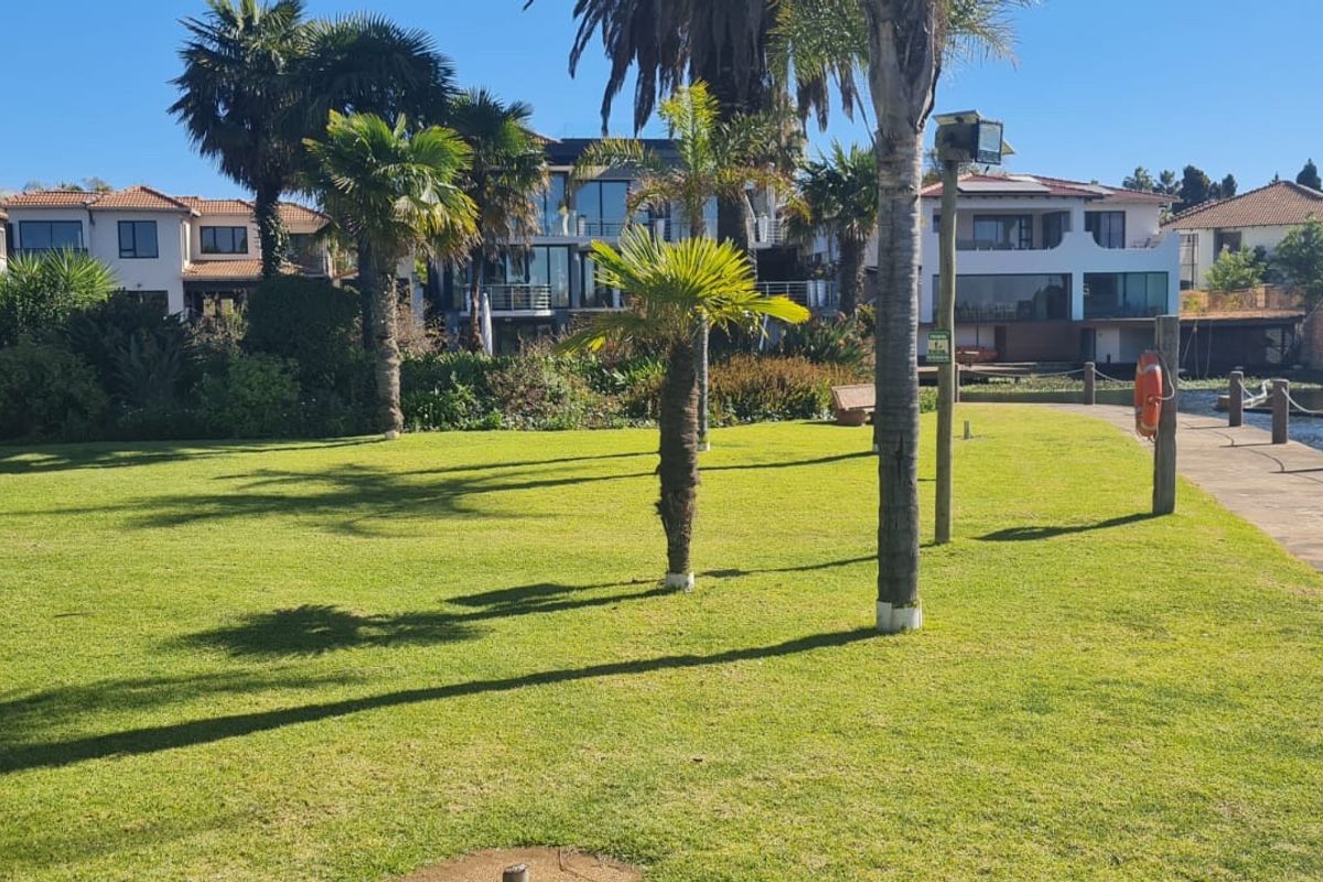 A well-maintained waterfront garden area featuring lush green lawns, mature palm trees, and a paved walkway along the canal edge.