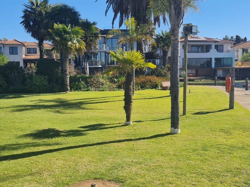 A well-maintained waterfront garden area featuring lush green lawns, mature palm trees, and a paved walkway along the canal edge.