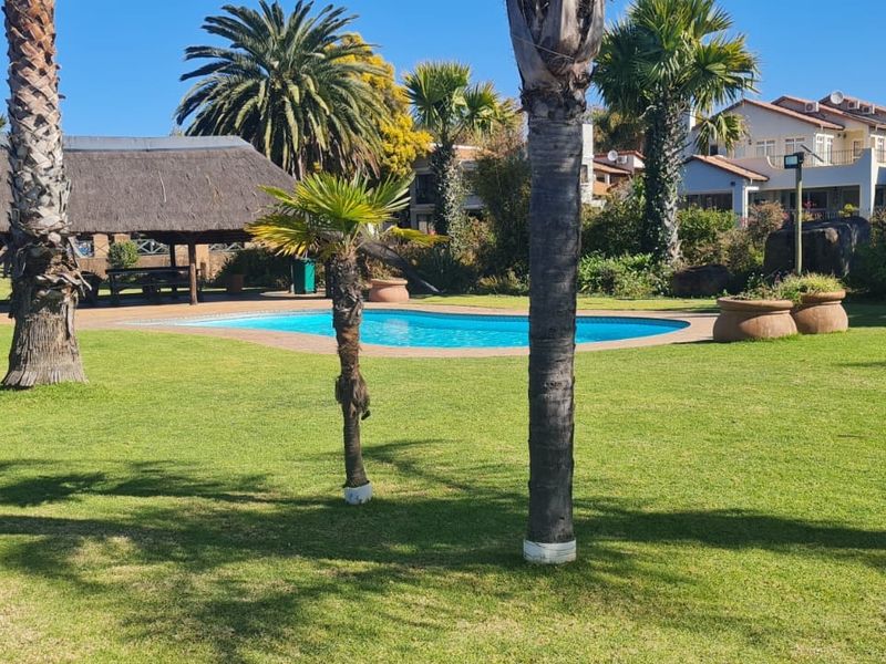 A well-maintained outdoor pool area featuring a swimming pool, a traditional thatched lapa, and established palm trees set within a spacious garden.
