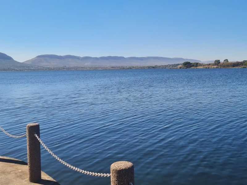 A scenic view overlooking a large lake with distant mountains, featuring a concrete pier edge with bollards and chain railing.