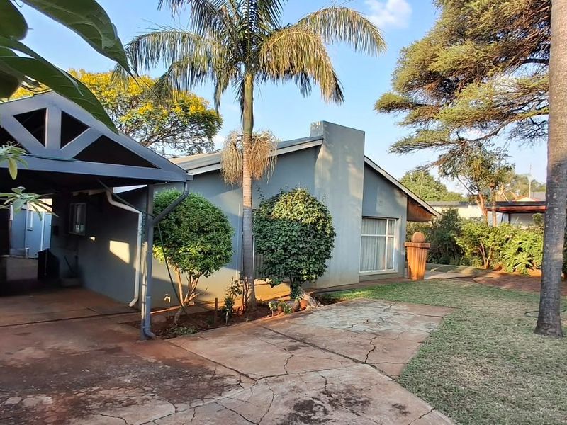 Front view of the property featuring a covered carport, paved driveway with visible cracking, and a mature landscaped garden.