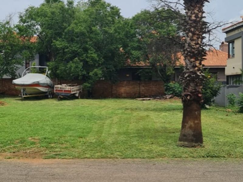 A vacant residential plot featuring a grass lawn, a mature palm tree, and a brick boundary wall, viewed from the street.