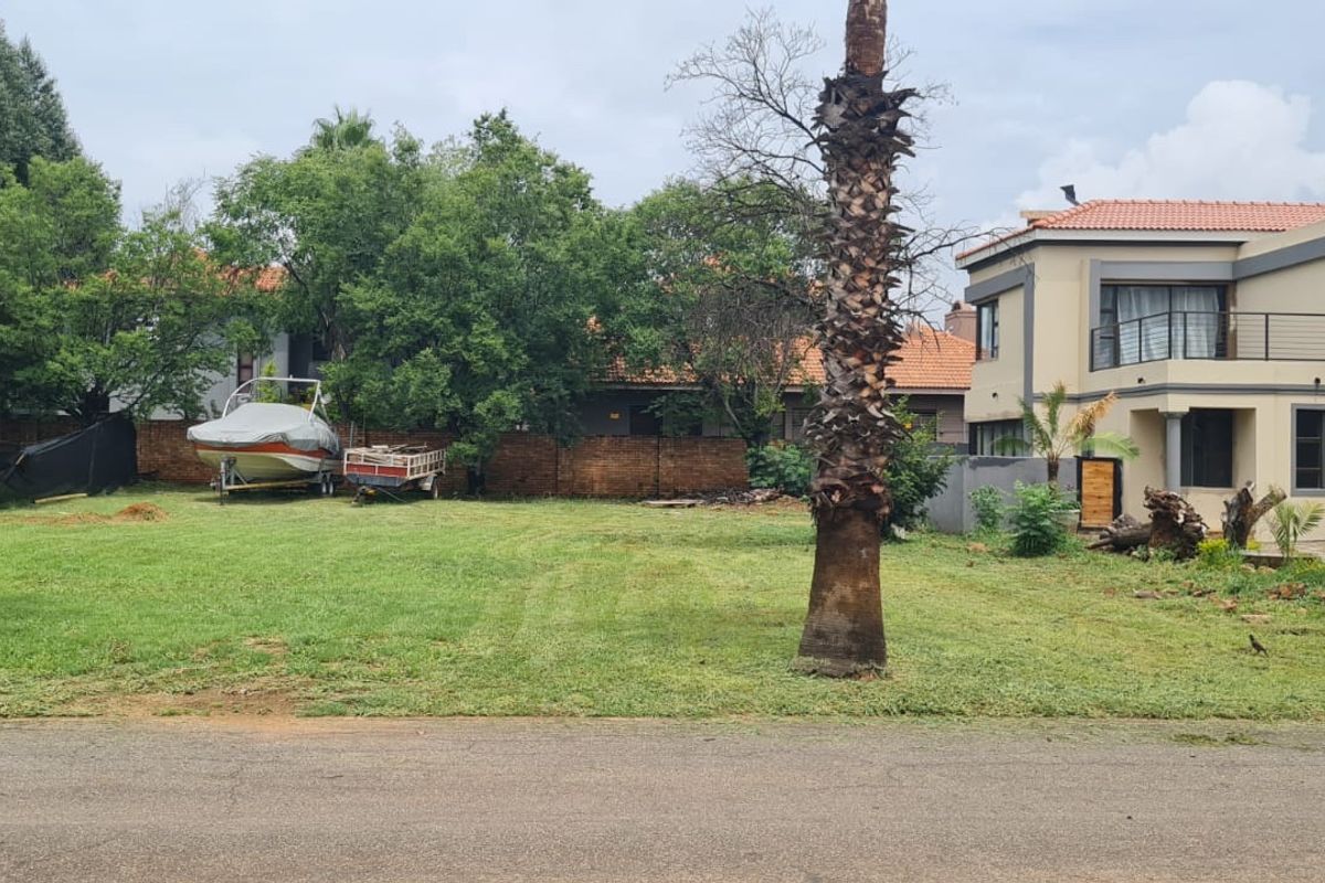 A view of a residential vacant land plot featuring a well-maintained lawn, mature palm trees, and a brick boundary wall.