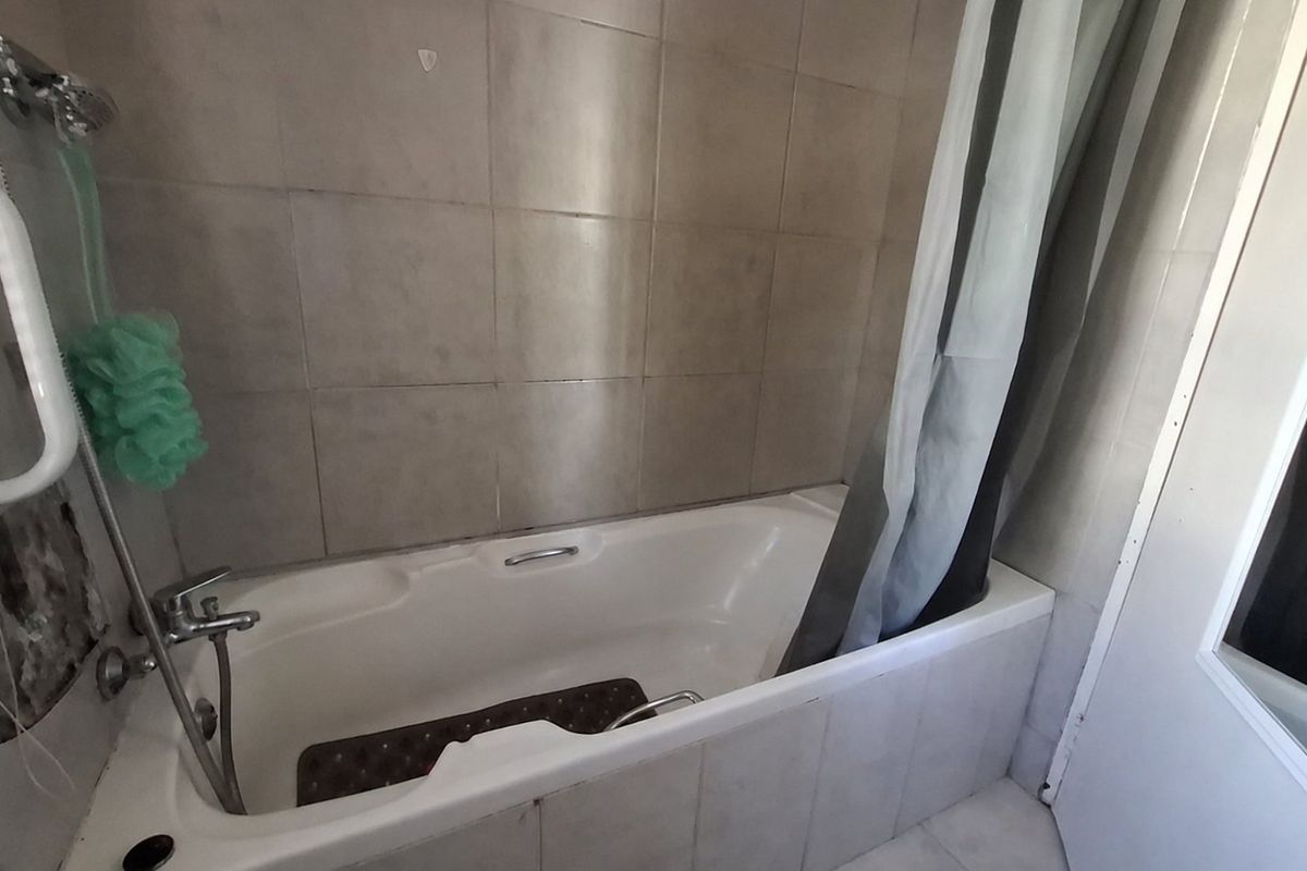 A bathroom featuring a bathtub with a shower mixer and wall tiling, showing visible wear and damage to the wall finish near the plumbing.