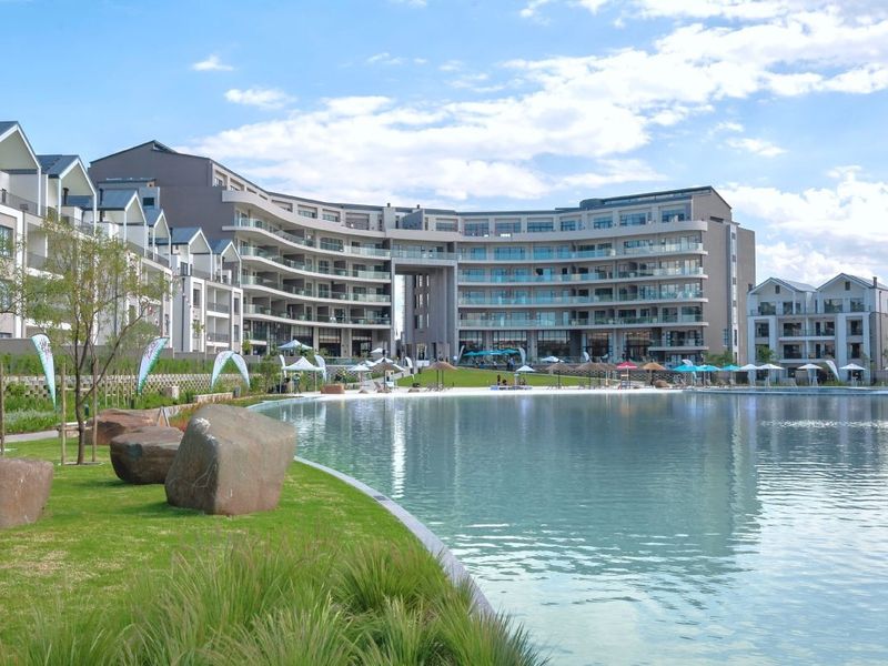 A scenic view of a large man-made lagoon surrounded by landscaped gardens and a modern multi-story residential apartment complex.