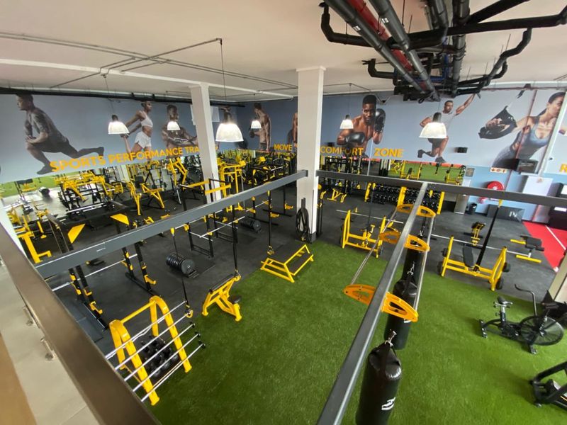 Spacious, well-equipped commercial-style gym featuring exposed industrial ceiling services, rubberized flooring, artificial turf zones, and extensive wall mirrors.