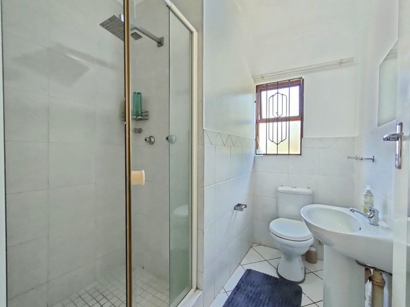 A functional bathroom featuring a shower enclosure, pedestal basin, and toilet with tiled finishes and a window with security bars.