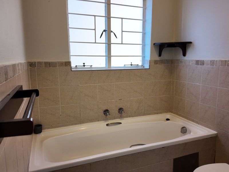A functional bathroom featuring a built-in bathtub with tiled splashback, security bars on the window, and wall-mounted fixtures.