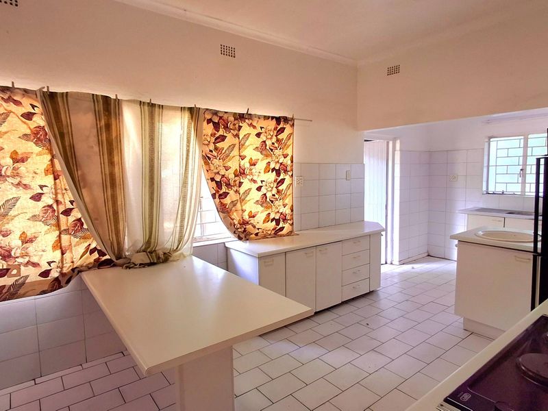 A spacious, dated kitchen featuring white tiled flooring, built-in cabinetry, a breakfast bar, and a tiled splashback, offering significant potential for modern renovation.