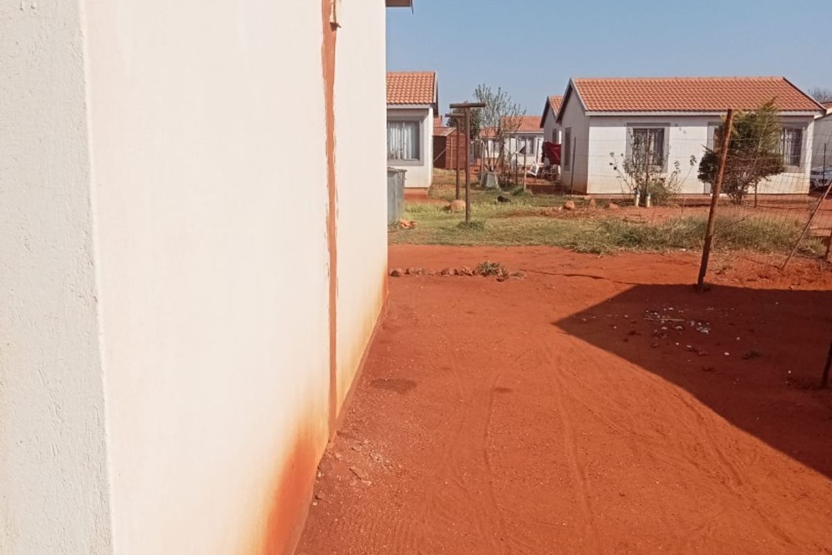 A view of the side and rear exterior of the house showing a plastered wall with visible staining and an unlandscaped dirt yard.