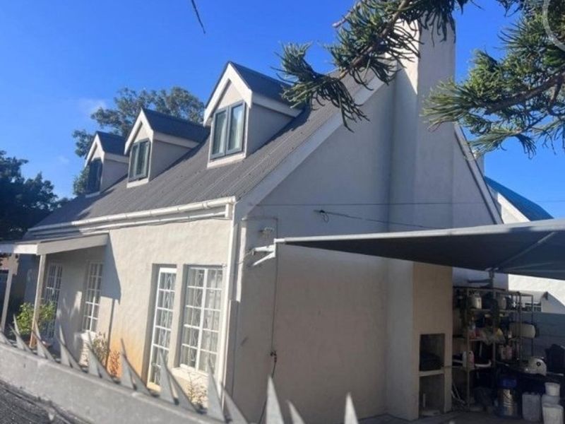 Rear view of a cottage-style house featuring a pitched roof with dormer windows, a chimney, and a built-in outdoor braai area with a shade sail.