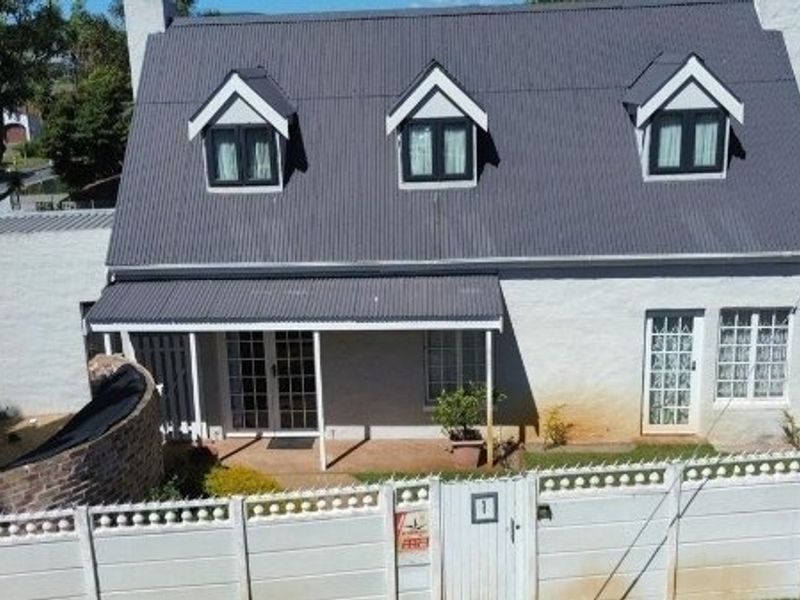 An elevated view of a charming cottage-style house featuring a corrugated metal roof, dormer windows, and a covered front patio behind a secure boundary wall.