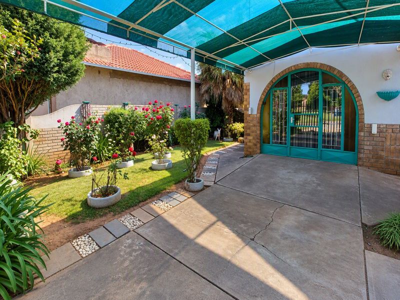 Covered patio area featuring concrete paving, an arched metal entrance door, and shade netting, adjacent to a landscaped garden with brick boundary walls.
