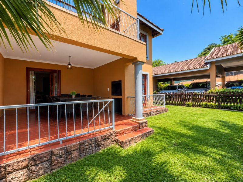 A spacious covered patio featuring terracotta tiling, stone cladding, and a built-in braai, overlooking a well-maintained garden and carport area.