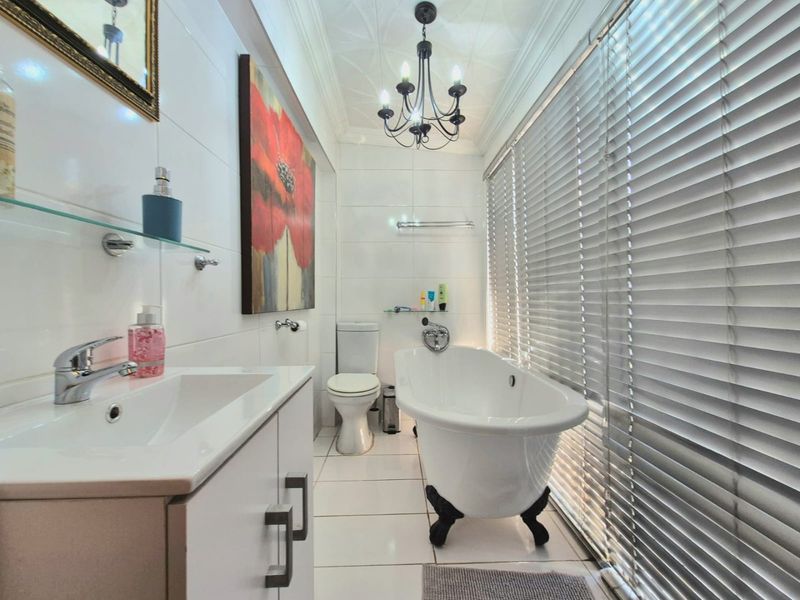 A bright, modern bathroom featuring a classic freestanding clawfoot bathtub, a vanity unit, and elegant chandelier lighting with large windows covered by venetian blinds.