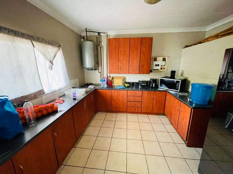 A functional kitchen featuring wooden cabinetry, tiled flooring, and ample counter space, though it would benefit from cosmetic updates and improved plumbing concealment.